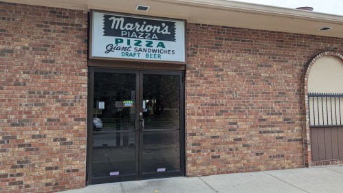 Exterior of Marion's Piazza, featuring a sign for pizza, giant sandwiches, and draft beer, with a brick facade.