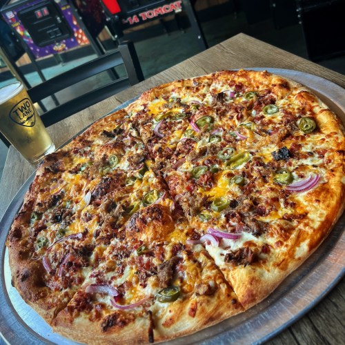 A large pizza topped with cheese, jalapeños, onions, and meat, served on a metal tray with a beer in the background.