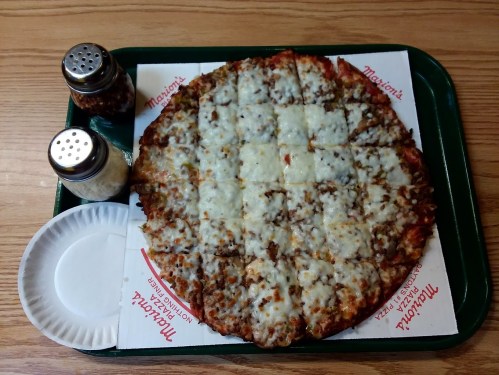 A round pizza with cheese and toppings, cut into squares, served on a tray with salt and pepper shakers.
