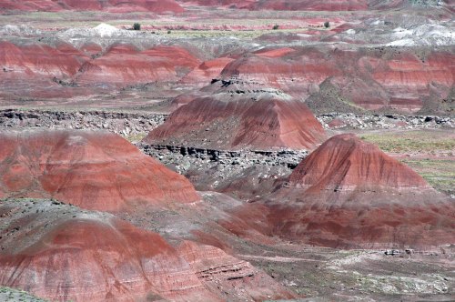 Colorful, layered hills and mesas in a desert landscape, showcasing shades of red, brown, and green under a clear sky.
