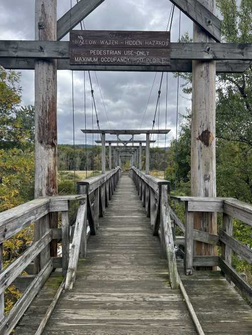 A wooden pedestrian bridge stretches over a river, surrounded by trees under a cloudy sky. Warning signs are visible.