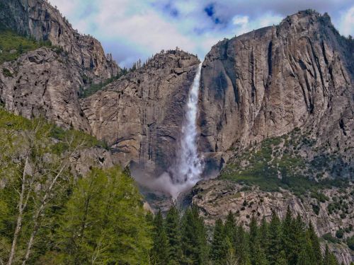A majestic waterfall cascading down a rocky cliff, surrounded by lush greenery and towering mountains under a cloudy sky.