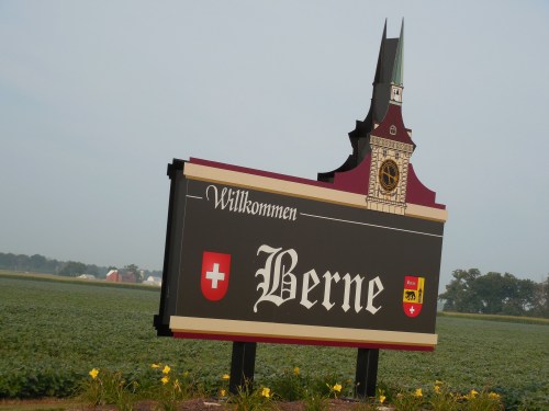 Welcome sign for Berne, featuring a clock tower design and Swiss flags, set against a rural landscape.