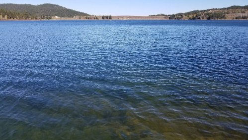 A serene lake with clear blue water, surrounded by green hills under a clear sky.