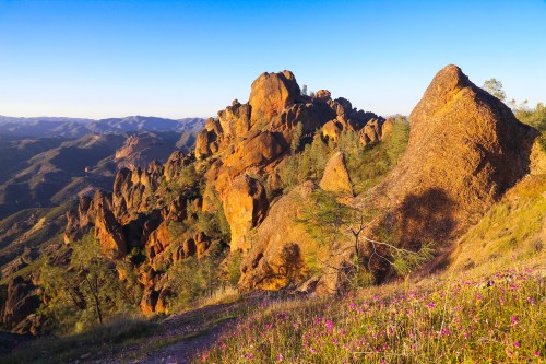 A scenic view of rugged orange rock formations surrounded by green hills and wildflowers under a clear blue sky.