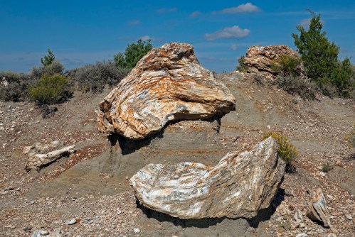 Two large, weathered rocks with layered textures sit on a dry, rocky landscape under a blue sky with scattered clouds.