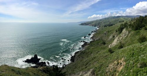 A panoramic view of a coastal landscape with green hills, rocky shores, and calm blue waters under a partly cloudy sky.
