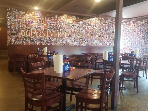A cozy restaurant interior with wooden tables, chairs, and a wall covered in photos, featuring the phrase "UGLY NEVER LOOKED SO GOOD."