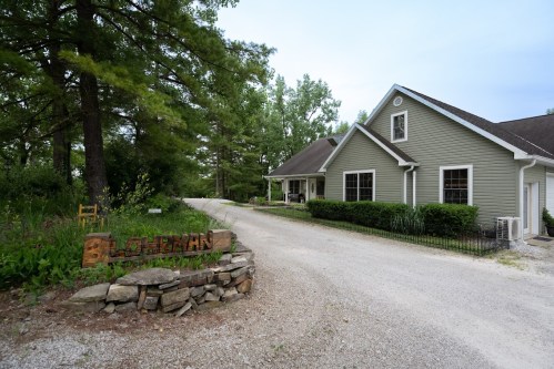 A gravel driveway leads to a green house surrounded by trees and shrubs, with a wooden sign reading "Lohman."