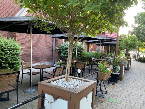 Outdoor dining area with tables, umbrellas, and planters along a brick wall, featuring greenery and flowers.