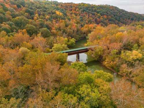 Aerial view of a bridge over a river, surrounded by vibrant autumn foliage in a lush landscape.