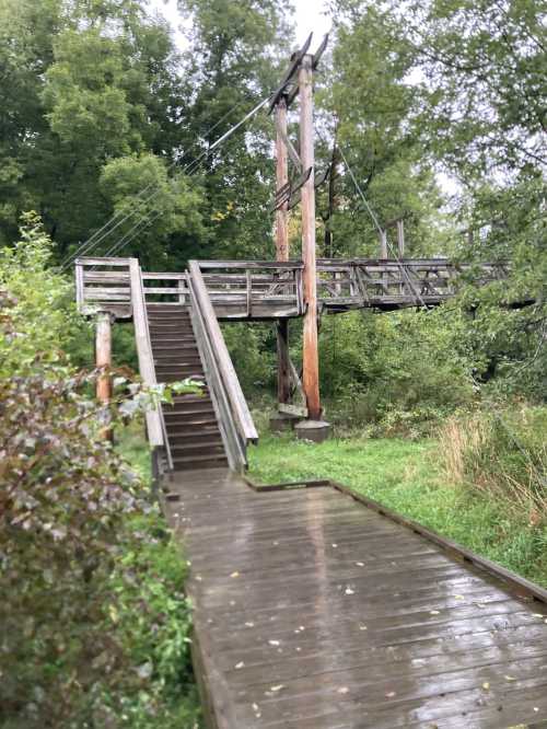 A wooden suspension bridge with stairs, surrounded by lush greenery and wet pathways.