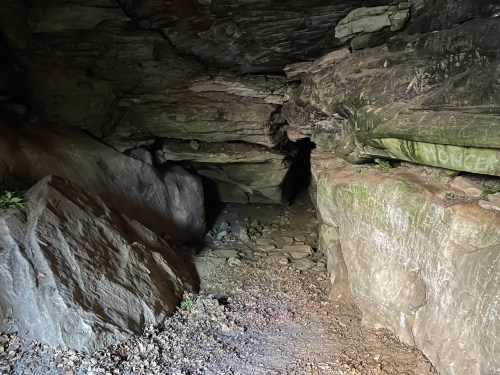 A dark cave entrance surrounded by large rocks and uneven terrain, with faint light illuminating the interior.