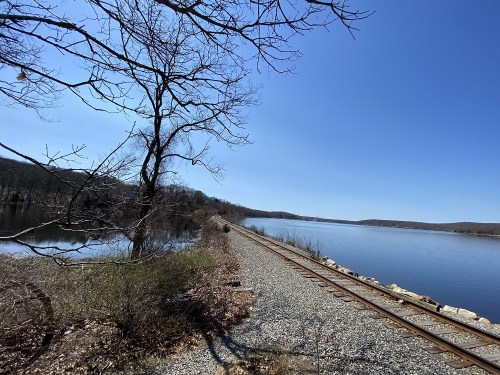 A serene landscape featuring a calm river, railway tracks, and bare trees under a clear blue sky.