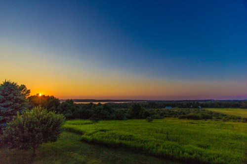 Sunset over a serene landscape with green fields and trees, reflecting warm colors in the sky.