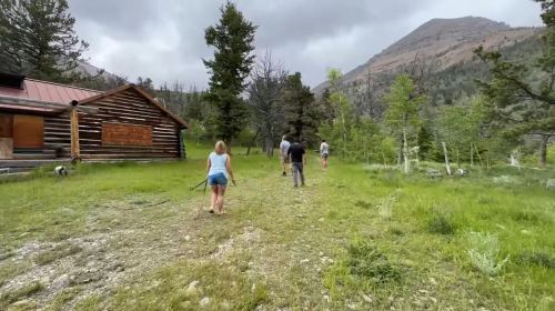 A group of people walking on a grassy path near a cabin in a mountainous forested area.