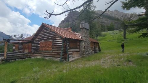 A rustic log cabin with a stone chimney, surrounded by green grass and mountains under a cloudy sky.