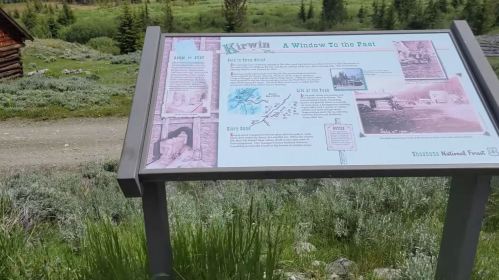 Sign about Kirwin, detailing its history and significance, surrounded by greenery and a dirt path in a scenic landscape.