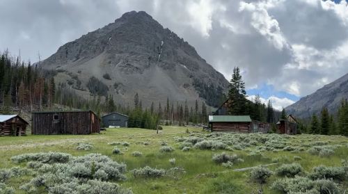 A scenic view of a mountain landscape with rustic cabins and lush green grass under a cloudy sky.