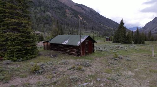 A rustic wooden cabin with a green roof, surrounded by trees and mountains in a serene, natural landscape.