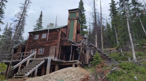 A rustic wooden cabin with a tall chimney, surrounded by tall trees and rocky terrain.
