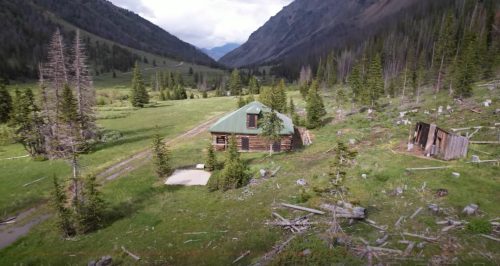 A rustic cabin with a green roof surrounded by trees and mountains in a lush valley.