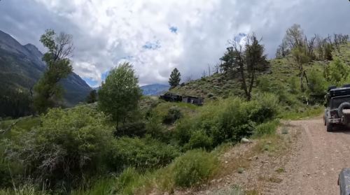 A rugged landscape with green vegetation, rocky terrain, and two off-road vehicles on a dirt road under a cloudy sky.