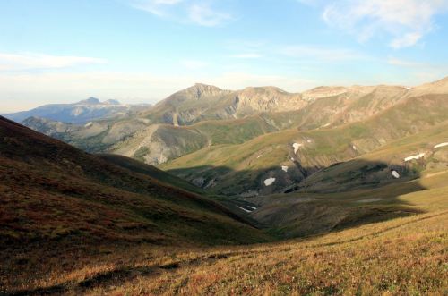 A scenic view of rolling mountains and valleys under a clear blue sky, with patches of snow and green grass.