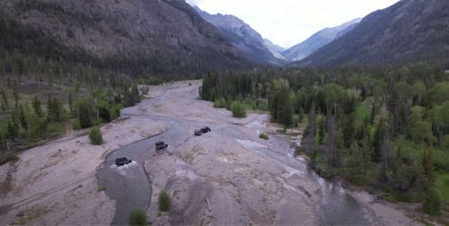 Aerial view of a rocky riverbed with four vehicles navigating through a lush valley surrounded by mountains.