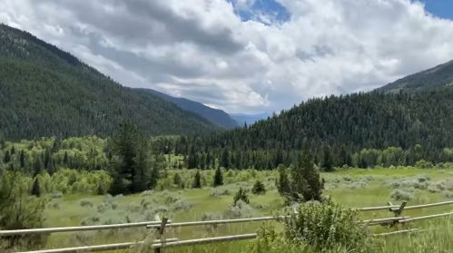 Lush green valley surrounded by mountains under a partly cloudy sky, with a wooden fence in the foreground.