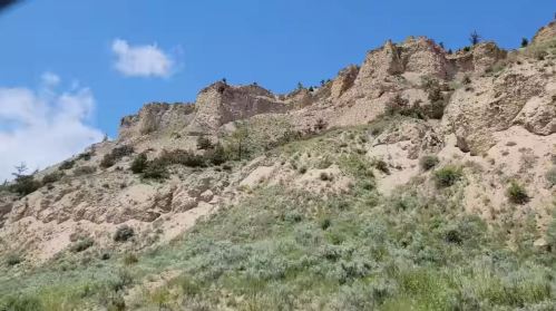A rocky hillside with steep cliffs and sparse vegetation under a bright blue sky with a few clouds.