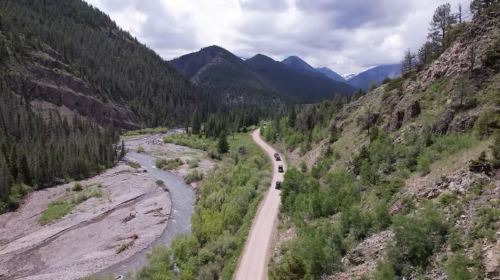 A winding dirt road alongside a river, surrounded by lush green trees and mountains under a cloudy sky.