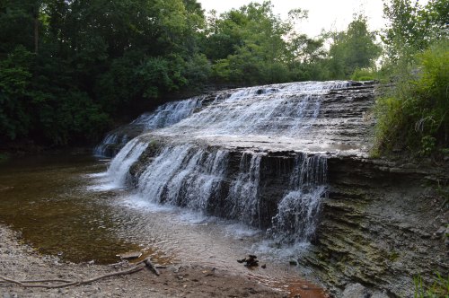 A serene waterfall cascading over rocky ledges into a calm stream, surrounded by lush greenery.