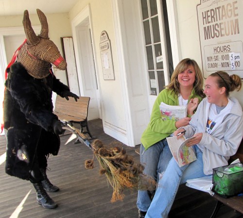A person in a horned mask playfully approaches two laughing women sitting on a bench outside a museum.
