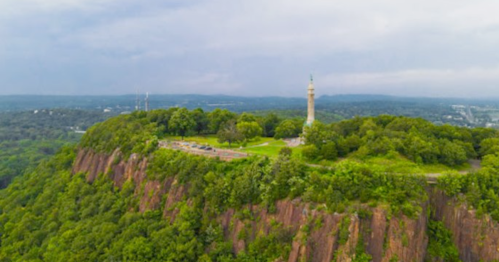 Aerial view of a green hilltop with a monument, surrounded by trees and a distant landscape under a cloudy sky.