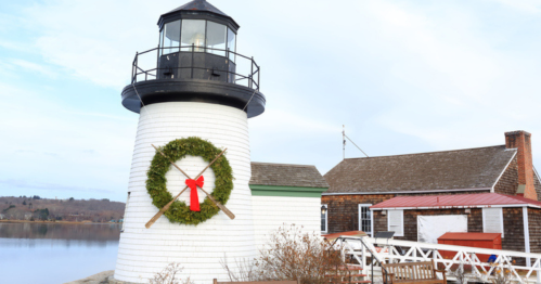 A white lighthouse adorned with a festive wreath and red bow, beside a calm water body and a rustic building.