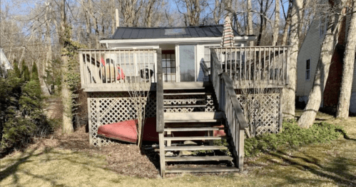 A small cabin with a deck and stairs, surrounded by trees and grass, featuring an American flag and a red couch.