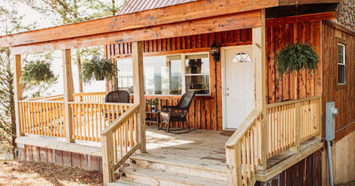 A cozy wooden cabin porch with two chairs, a small table, and hanging plants, surrounded by trees.