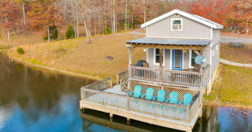 A cozy cabin by a pond, featuring a deck with blue chairs and surrounded by autumn foliage.