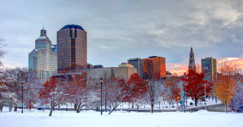 City skyline with modern buildings, autumn trees, and a snowy foreground under a cloudy sky at sunset.