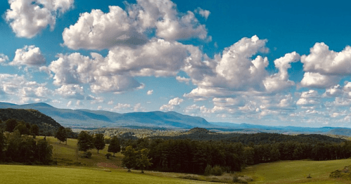 A panoramic view of rolling hills and mountains under a bright blue sky filled with fluffy white clouds.