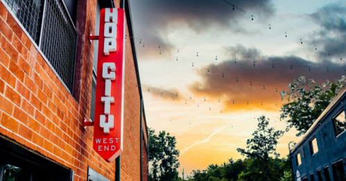 A vibrant sunset behind a building with a red "Hop City West End" sign and string lights overhead.