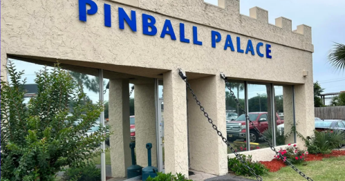 Exterior of Pinball Palace, featuring a castle-like design and blue signage, surrounded by greenery and parked cars.