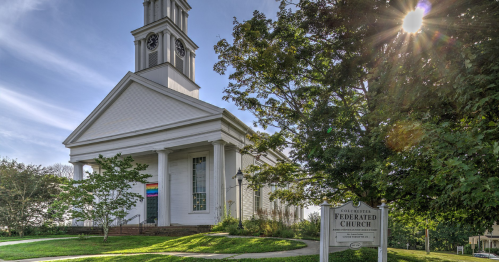 A white church with a clock tower, surrounded by trees and a sign reading "Federated Church." Sunlight shines in the background.
