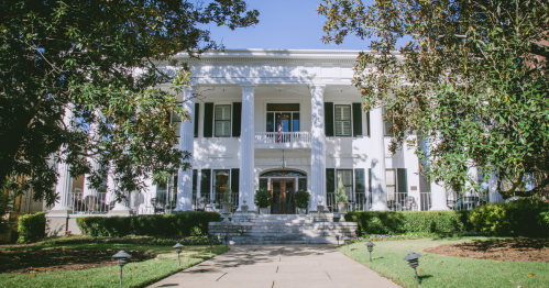 A grand white mansion with columns, surrounded by greenery and a clear blue sky. Steps lead to the entrance.