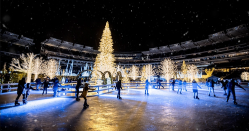 A festive ice skating rink surrounded by illuminated trees and a tall Christmas tree, set in a stadium at night.