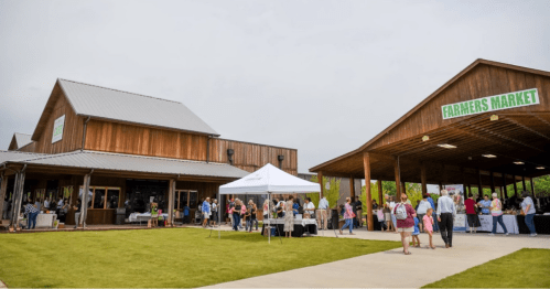 A bustling farmers market with people browsing stalls outside wooden buildings under a cloudy sky.