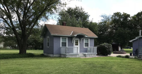A small purple house with a front porch, surrounded by green grass and trees under a partly cloudy sky.