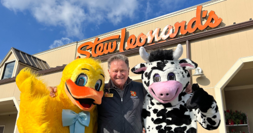 A man poses with a duck and cow mascot outside a Stew Leonard's store under a blue sky.
