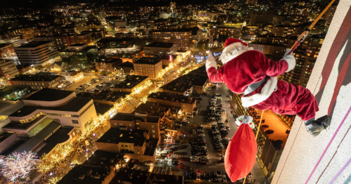 Santa Claus rappelling down a building at night, overlooking a city adorned with festive lights and decorations.